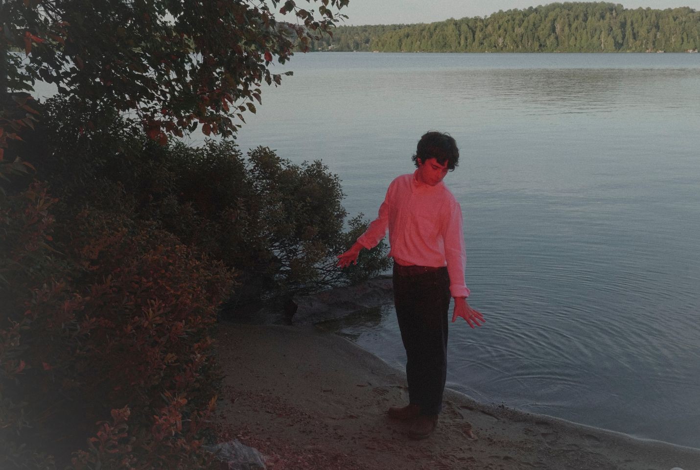 A Man wearing a white long sleeve top, black trousers, brown belt, and brown shows, with curly dark brown hair stands at the edge of a lake on the sand with his back to the water. There is a red hued light shining towards him